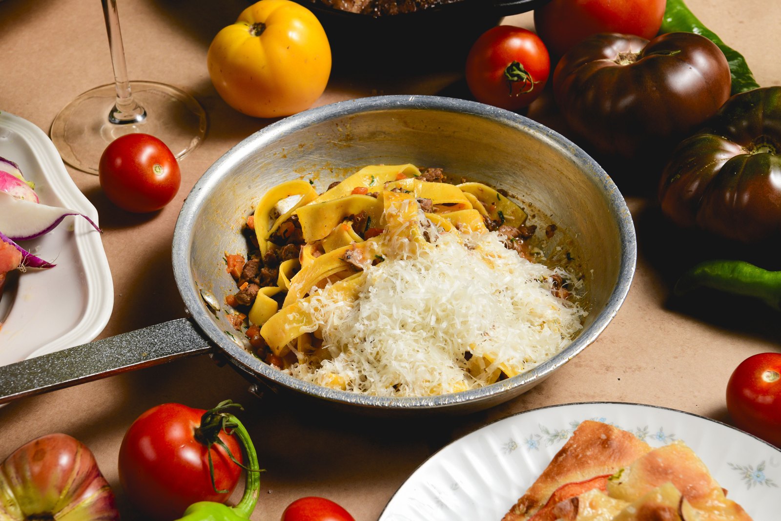 Lamb ragù pasta in a metal pan with grated cheese, surrounded by tomatoes
